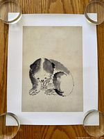 Top-down photo of sumi-e ink wash art print showing a fluffy puppy on textured washi paper with wide margins on a wood surface, held flat by four clear weights.