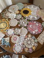 Collection of multicolored and white crochet doilies on table, showcasing diverse shapes including round, oval, and novelty floral designs with various colors and embroidery.