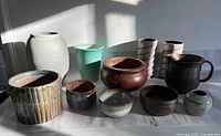 Photo showing the assorted 11 studio pottery pieces including vases, pitcher, round planter, ribbed vessel, and bowls displayed on a table with strong natural light.