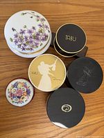 Top-down photo of six vintage powder and trinket boxes including Dana Tabu boxes, Limoge floral porcelain boxes, and a black monogrammed box.