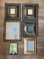 Top-down view of all seven frames laying flat on wooden surface. Includes three matching dark baroque square frames with deep relief scrollwork, one larger ornate gold-tone frame, one gilded simple frame, one small framed colorful quote print with art deco style illustration, and backing for one frame.