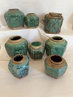 Front view of 8 green glazed vintage Chinese stoneware jars showing various shapes and sizes with hand-pressed relief motifs and cordage wrapping on one jar.