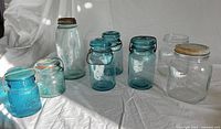Eight vintage glass storage jars arranged on white cloth including aqua and clear glass jars with various types of lids.