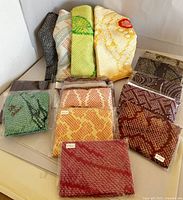 Overview of 15 pieces of shibori silk textiles in various colors and patterns displayed folded and some in plastic packaging.