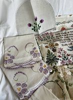 Photo showing handmade embroidered linens including a purple floral embroidered cloth with scalloped edges and a small oval doily embroidered with purple flowers