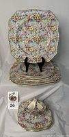 Stack of square plates with floral patterns and scalloped edges, some stacked against a white backdrop with a display stand.