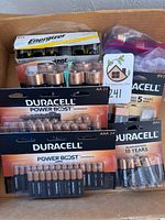 Photo showing multiple unopened packs of batteries in a cardboard box. Visible brands include Duracell and Energizer. Sizes include AA, AAA, C, D and 9V. Packaging shows 'Power Boost' line for some Duracell batteries.