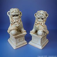 Pair of ivory-toned glazed ceramic Foo Dogs standing on rectangular stepped plinth bases with one paw on a textured sphere, shown against a blue backdrop