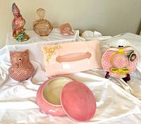 Seven pink-toned decorative items displayed on white fabric including ceramic kitten yarn holder, cockatoo figurine, pink tissue holder, sunhat wall pocket, ceramic jar with lid, crystal trinket box, and glass paperweight.