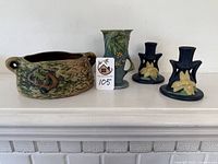 Front view of all four pottery pieces: round handled casserole, small vase with floral relief and cutout handles, pair of matching candlesticks with floral decoration.
