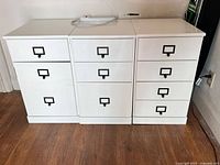 Front view of three white cabinets side by side showing different drawer configurations and black metal label pulls.