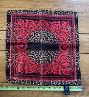 Full view of the Versace silk scarf laid flat showing the baroque red pattern over leopard print background. Measuring tape shows approximate size of 16 inches square.