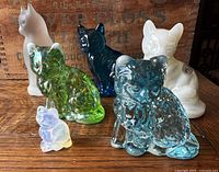 Overview of six cat figurines showing different colors, materials, and finishes including iridescent green, blue, white opalescent, cobalt, and milky stone-like.