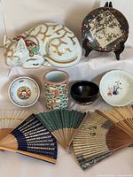 Front view of all 11 vintage Asian decorative pieces arranged including ceramic sleeping cat, decorative plate on stand, assorted bowls, pink and green brush pot, black lacquerware bowl, celadon dish, and six folding fans in various styles and colors.