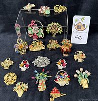 Wide shot showing 20 vintage flower basket brooches with colorful rhinestones, pearls, and enamel on a black background, arranged in multiple rows and a clear display stand.