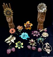 Full lot of floral and figural fruit brooches displayed on black background with two wooden hand holders showing sunflower and white flower brooches