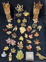 Full lot of 35 vintage leaf-themed brooches displayed against a black fabric background, featuring various colors, materials, and sizes including enameled and gold-tone pieces.