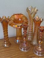 Front view showing four ruffled-edge marigold carnival glass vases and two candlesticks on wooden surface against plain wall.