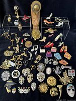 Full overhead view of lot showing variety of vintage brooches with figural and novelty themes including shoes, hats, masks, music notes, handbags, and cameos arranged on black background.