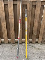Two long extension poles leaning against a wooden fence showing overall condition and length