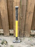 Full view of the Black + Decker 8lb sledge hammer leaning against a wooden fence, showing yellow handle with black rubber grips and metal hammer head.