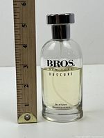 Clear glass bottle of BROS New York Obscure cologne with silver cap next to ruler showing approx 5 inch height.