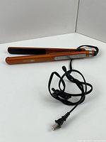 Hair straightening iron shown with power cord, orange metallic finish, and black ceramic plates.