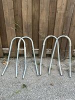 Four metal stand-off arms for extension ladder, U-shaped tubular design, arranged outdoors against wooden fence.