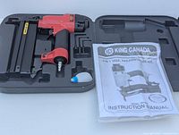 Red and black pneumatic nailer/stapler with oil bottle, hex keys and instruction manual in a plastic carrying case.