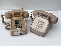 Two vintage telephones side by side on a white surface. Beige phone with black keys to the left; tan phone with cream keys to the right.