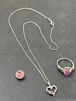 Photo showing a sterling silver chain necklace with a heart pendant featuring a small pink stone, a pink bead charm, and a sterling silver ring with a pink heart-shaped gemstone with crown detail.