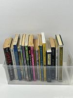 Photo showing 15 assorted books in various conditions arranged inside a clear plastic bin, some paperbacks with creased spines and some hardcover.