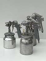 Three metallic spray guns with attached canisters, viewed from a slight angle showing front and side of the items.