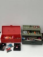 Two fishing tackle boxes, red and dark green, with contents visible inside including fishing floats, hooks, sinkers, and fishing line.