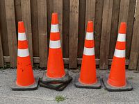 Four large orange traffic pylons with black square bases and white reflective stripes arranged in a row against a wooden fence, showing signs of use.