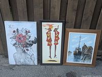 Three framed paintings displayed against a wooden fence, including a floral still life, a stylized figures painting, and a maritime scene.