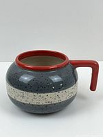 Side view of ceramic mug shaped like a curling stone with speckled gray body, white stripe, and red handle.