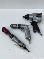 Photo showing three air power tools: pneumatic ratchet wrench with red head, pneumatic drill with chuck, and impact wrench with rubber grip.