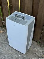Front side view of the white and gray Noma dehumidifier unit standing on ground with fence background.