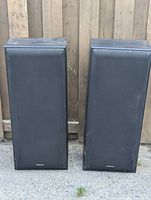 Pair of large Technics speakers standing side by side outdoors against a wood fence, showing fabric grill fronts.