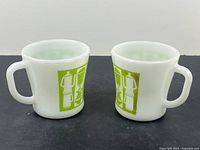 Pair of white opaque glass mugs with green graphic kitchen-themed designs on side, wide handles visible, resting on black surface.