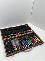 Top view of the open wooden art kit showing colored pencils on upper half and markers, watercolors, pastels, and accessories on lower half.