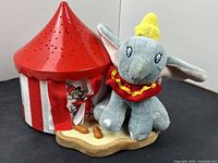 Red and white circus tent styled night light with perforations for star and moon shapes next to a plush Dumbo with yellow hat and red collar, sitting on beige base.