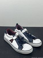 Pair of youth casual running shoes placed side by side on black surface showing front and outside sides. White with navy and maroon details, Velcro strap included.