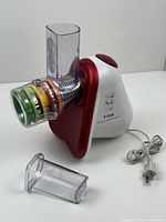 Full view of T-Fal red and white electric vegetable spiralizer with clear plastic feeding chute and power cord; includes small clear plastic food pusher attachment lying next to unit.