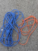 Two outdoor extension cords, one blue and one orange, lying coiled on a paved surface showing condition and length approximately.
