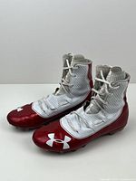 Front and side view of the pair of Under Armour high top soccer cleats, showing white and red coloration with logo on toe.