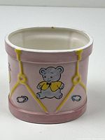 Oval ceramic vase with painted teddy bear, baby banner, and booties on pink base with yellow accents, showing front and side views.