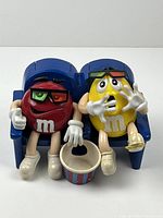 Front view of Red and Yellow M&M characters seated in blue theater chair with popcorn bucket in front.