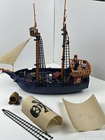 Side view of vintage Playmobil pirate ship with two tall orange masts, navy blue hull, and rigging ladders. Three pirate figures on deck. Two sails detached in front beside a brown cannon and rigging piece.